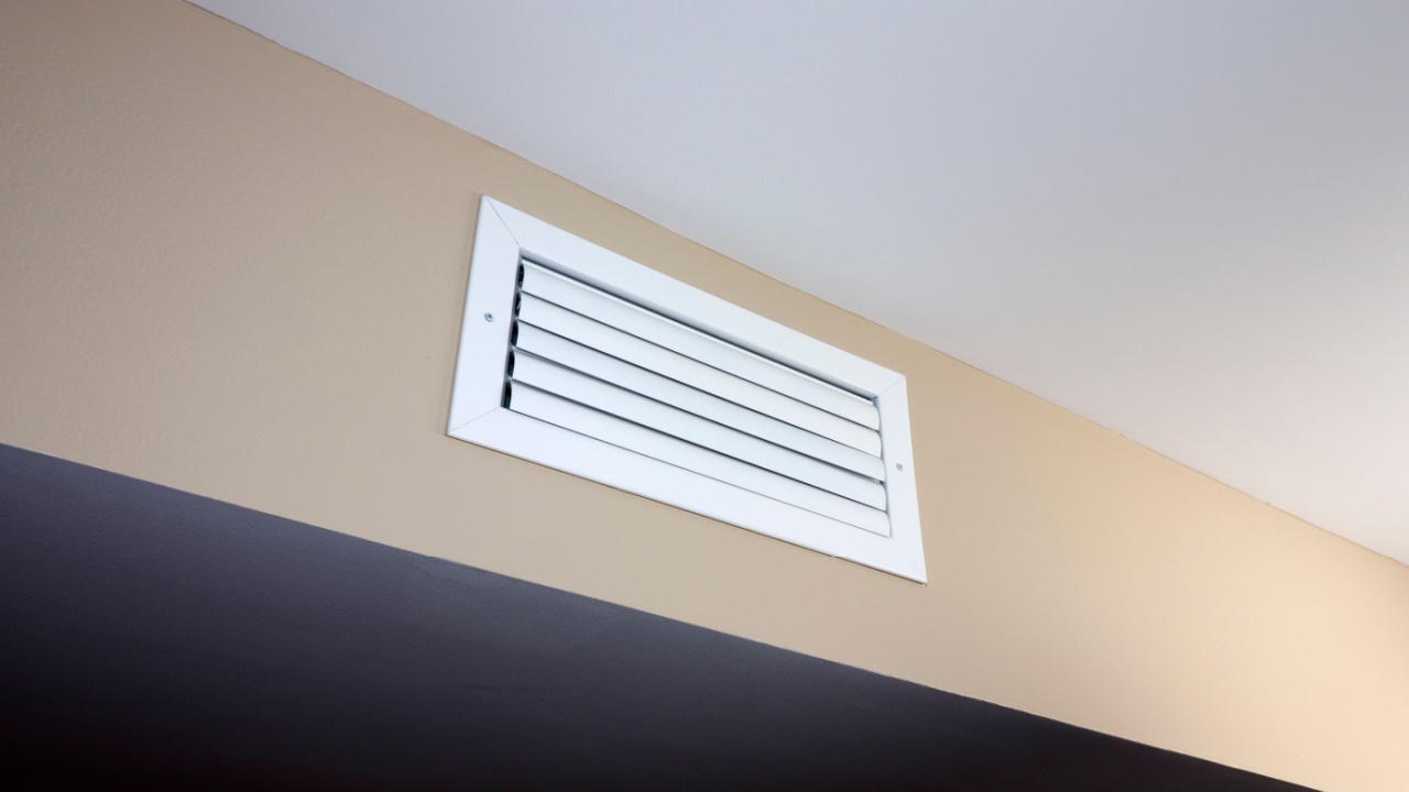 A white rectangle heating and cooling outflow air vent from a duct of an indoor HVAC system in a beige color wall near a ceiling. A small white rectangle outflow air duct vent inside a modern home.