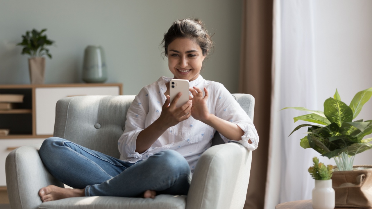 Happy pretty millennial Indian girl relaxing at home, resting in armchair, typing on smartphone, using online app, software, shopping on Internet, making video call. Mobile phone communication