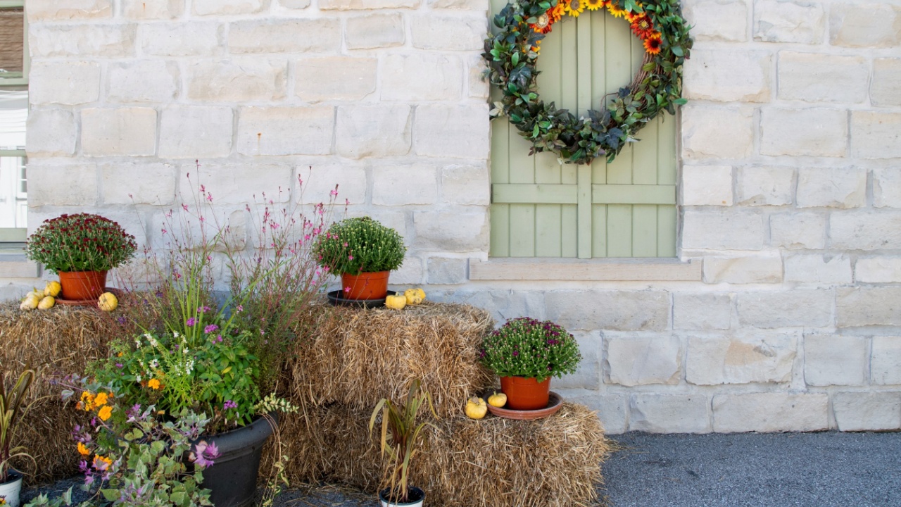 Fall DIY hand crafted porch decor with an autumn wreath, hay bales, mums, and flowers against a stone wall and green-shuttered window