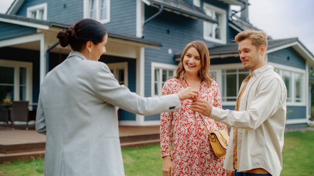 Happy Real Estate Agent Congratulating Young Homeowners with Their New Home Acquisition. Cheerful Couple Shaking Hands with the Businesswoman and Receiving Keys From Their Property.