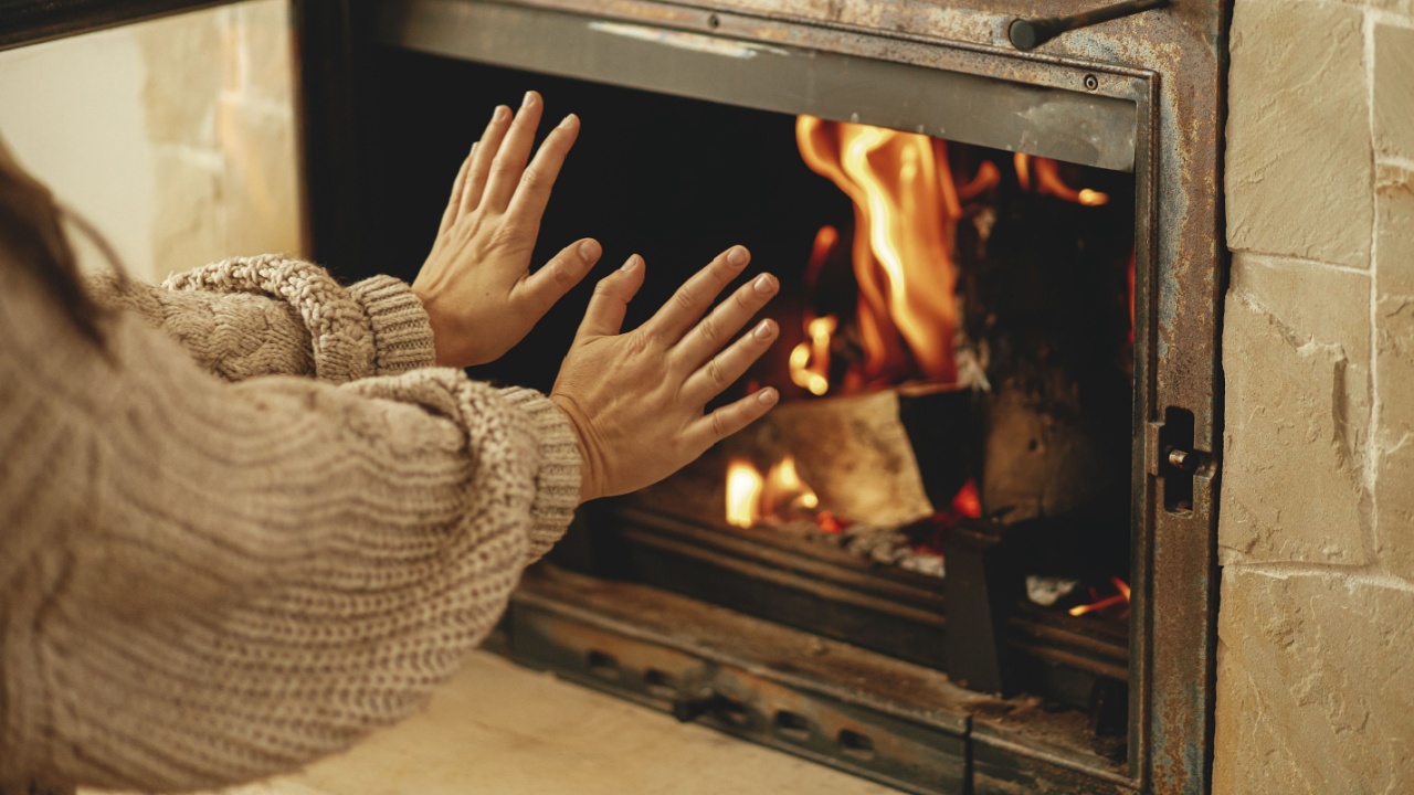 Heating house in winter with wood burning stove. Woman warming up hands at burning fireplace in rustic room in farmhouse. Fireplace heating alternative to gas and electricity