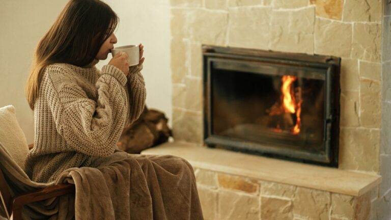 Woman in cozy sweater drinking cup of warm tea at fireplace in rustic room. Heating house in winter with wood burning stove. Young stylish female relaxing at fireplace in farmhouse