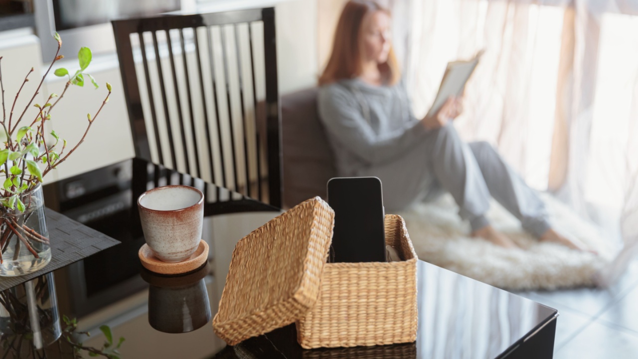 Smartphone is in separate wicker box on table. Woman reading a book in background. Stop using digital gadgets at home in favor of reading books and meditation. Mental and digital detox concept