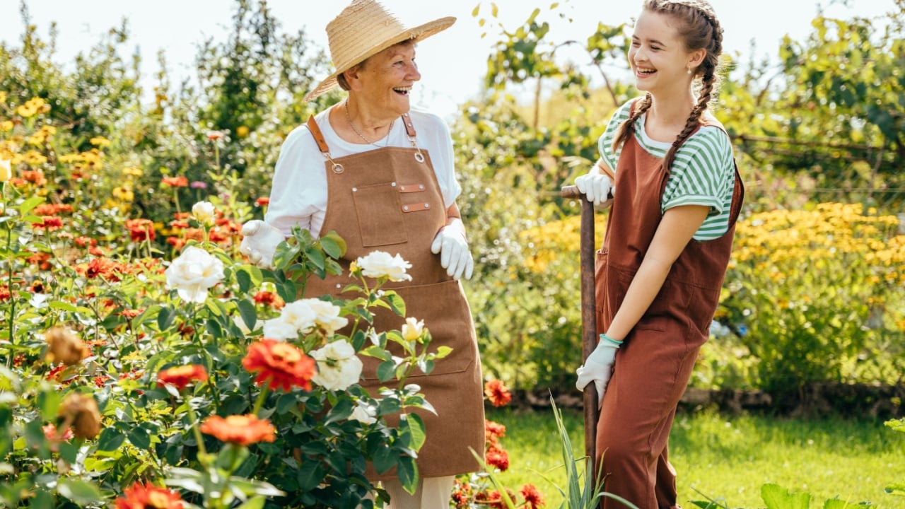 Portrait of teenager granddaughter with senior grandmother laughing duringwork in garden, with flowers around.