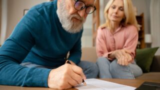 Senior Couple Filling In Form Signing Papers Sitting On Sofa Near Table Indoors. Paperwork Concept. Elderly Man Writing His Will In Notary Office. Cropped Shot, Selective Focus