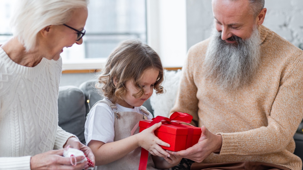 Attractive senior grandparents exchanging Christmas and New Year gift box with granddaughter, cute little girl. Looking happy and excited sitting near decorated tree in red Santa hats