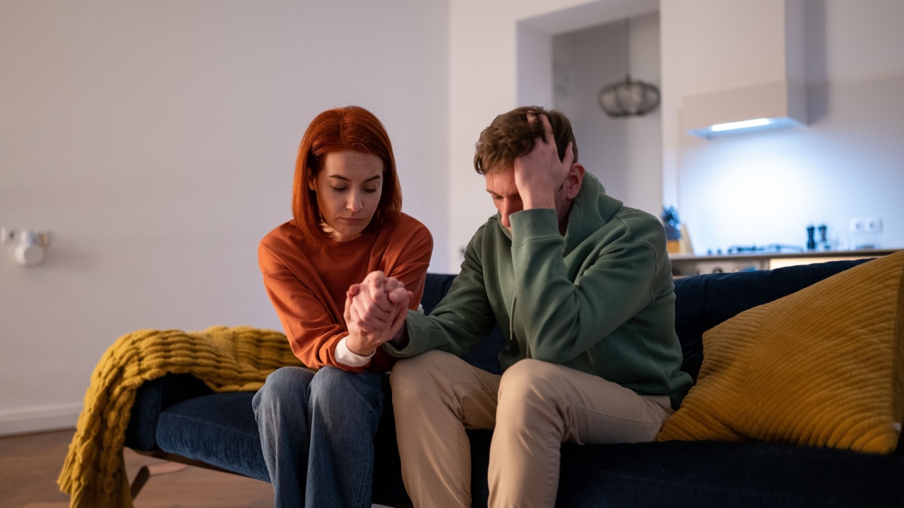 Young loving supportive woman wife holding hand of worried husband, showing support for partner during difficult times. Upset unhappy family couple holding hands while sitting together on sofa at home