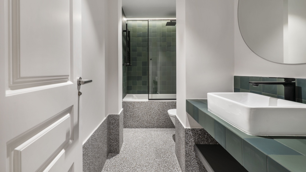 a modern design bathroom with a bathtub with a tempered glass screen, green tiles, porcelain sinks, a circular mirror, white carpentry, black details and gray terrazzo floors