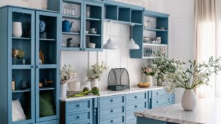 Vintage kitchen interior. Blue wooden cupboard with glass doors, storage drawers and chic decorative elements. Marble kitchen island with vase and flowers. Tableware on wooden shelves.