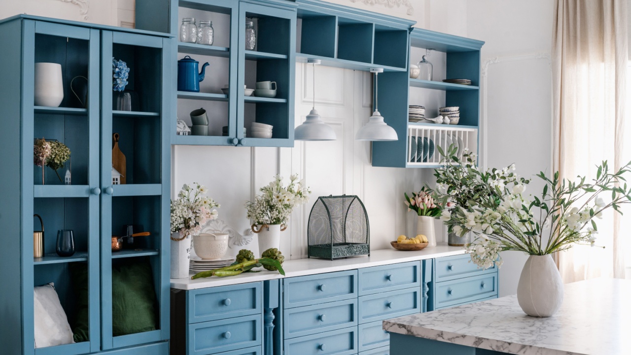 Vintage kitchen interior. Blue wooden cupboard with glass doors, storage drawers and chic decorative elements. Marble kitchen island with vase and flowers. Tableware on wooden shelves.