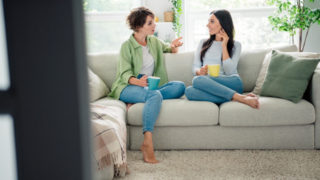 Full body portrait of two pretty cheerful lady barefoot sit comfy sofa drink coffee communicate dialogue pastime apartment indoors