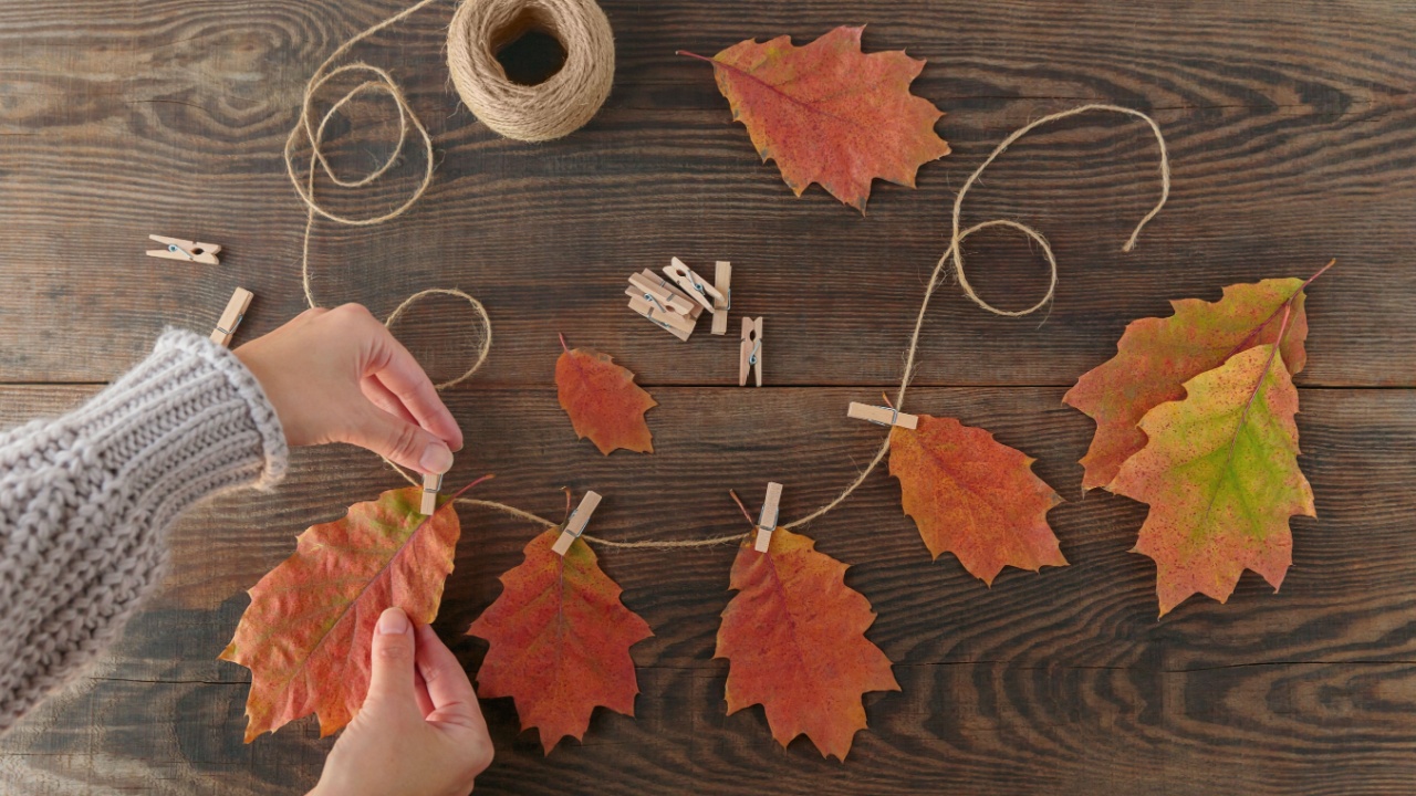 Homemade garland of colored autumn leaves. Womans hands hanging leaves on rope with clothespin on wooden background. Home diy decor for celebrating fall holidays, Thanksgiving and Halloween