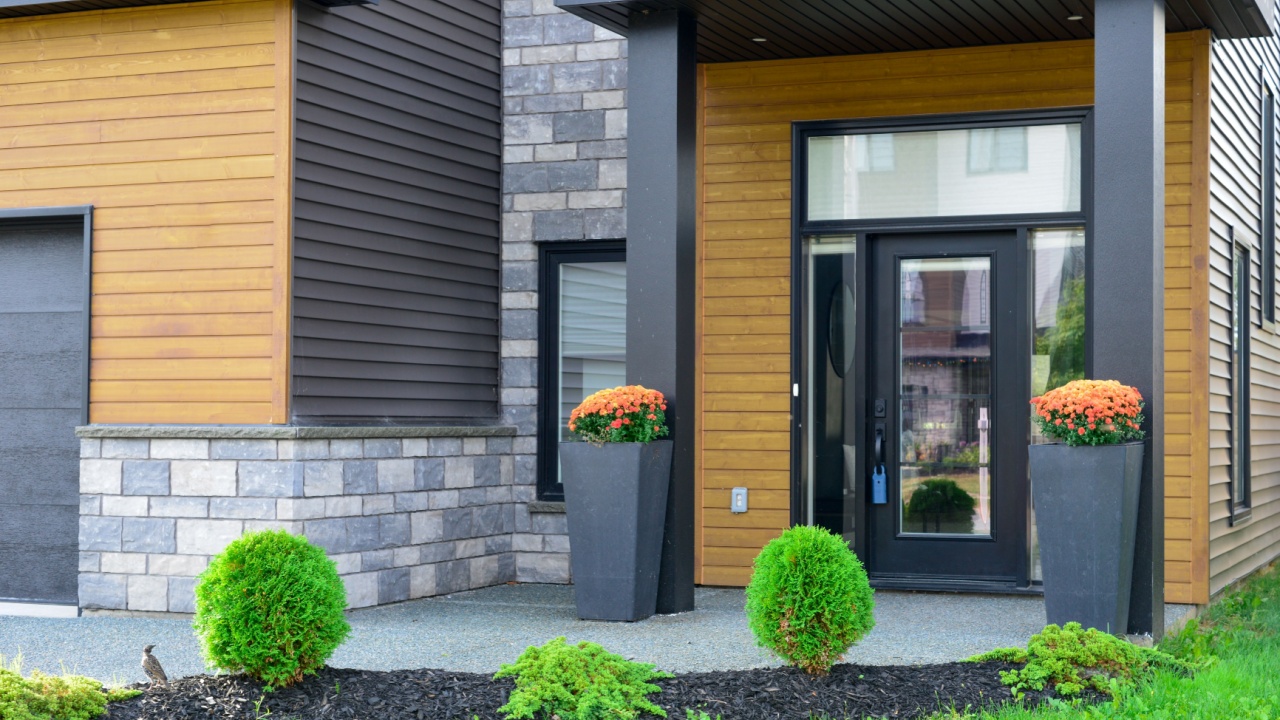 A street view of a modern suburban style executive house. There's a concrete walkway, the exterior walls are brown wood siding and grey brick, and the garage door is a large solid black metal door.