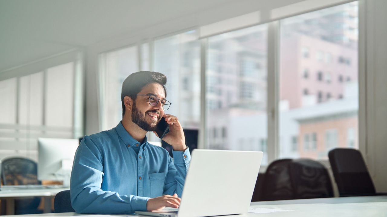Young happy busy latin businessman talking on phone using laptop computer in office. Smiling hispanic business man making call on mobile cellphone, consulting client or having work cell conversation.