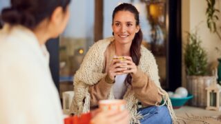 A smiling woman talking with her friend, sitting outdoors during the autumn season, holding a cup of coffee.