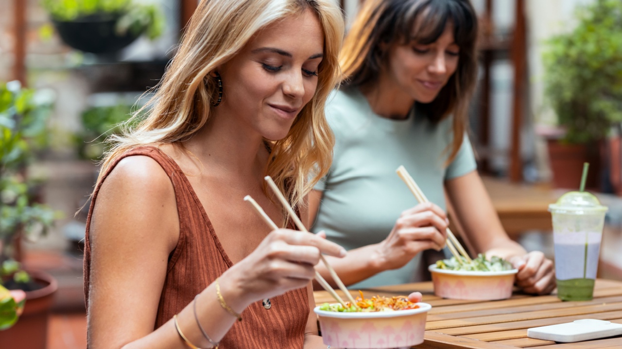 Shot of two smiling beautiful women friends eating pokes with chopsticks while talking in the restaurant terrace.