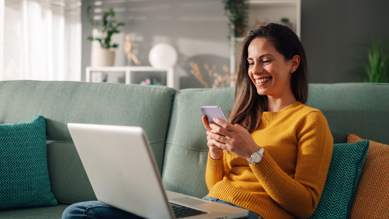 Portrait of a woman using laptop and a phone and checking email or news online while sitting on sofa at home. Searching for friends in internet social networks or working on computer. Copy space.