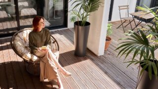 Smiling young professional business woman with red hair holding mobile cell phone in hands relaxing at work sitting in chair in sunny office looking away using smartphone