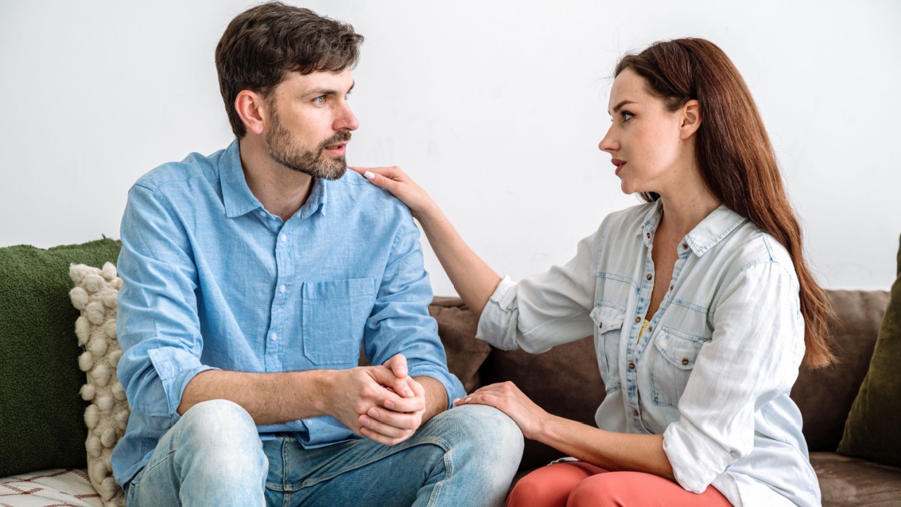 Support, trust and understanding in marriage. Young husband sitting on sofa at home and talking about problems, wife listening carefully and hugging man. Overcome family difficulties together.