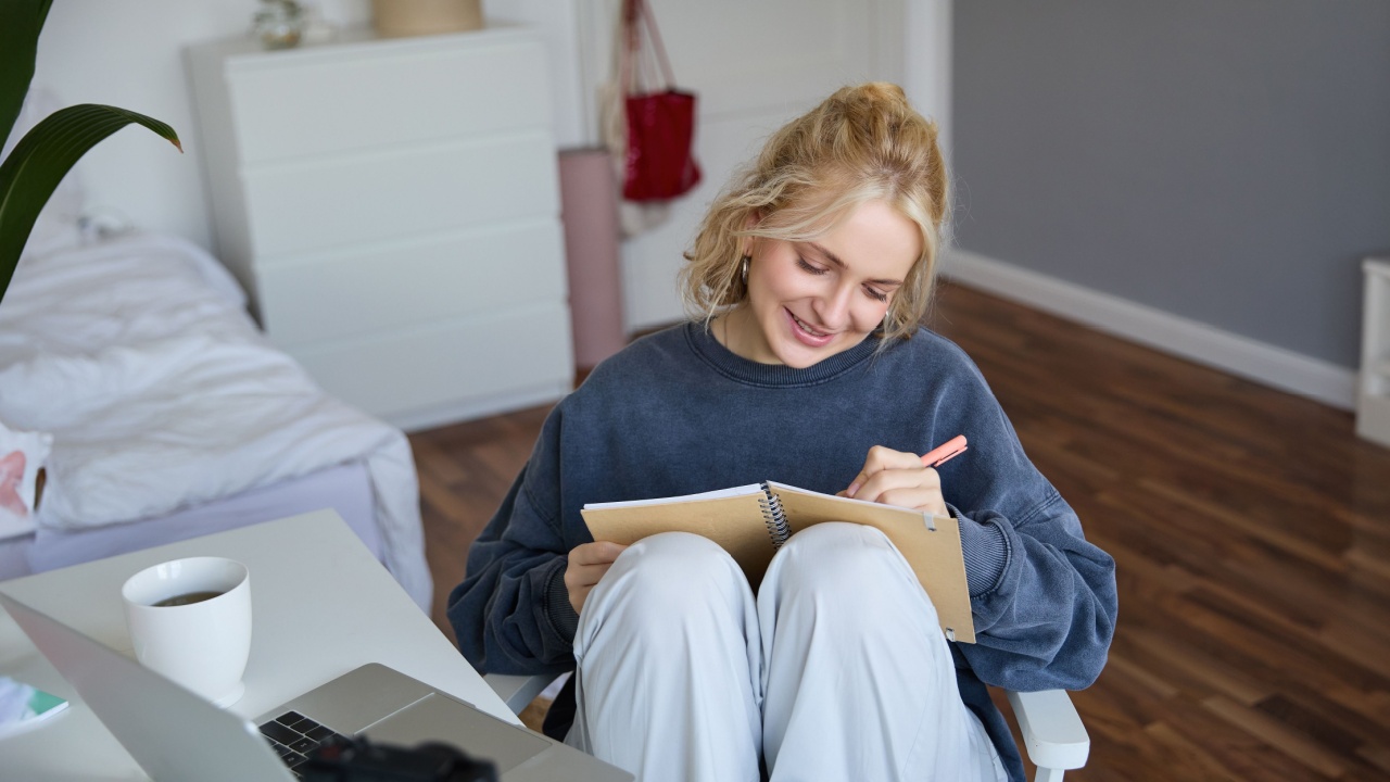 Portrait of smiling cute woman, lifestyle blogger, sits in her room with daily journal or planner, records video on digital camera, creates content for social media about daily routine.
