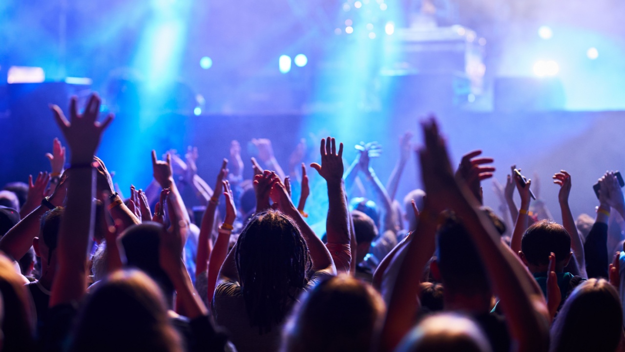 Concertgoers raise hands in rhythm at outdoor music festival, colorful stage lights illuminate excited audience. Fans enjoy live performance, nightlife energy, summer event brings people together.