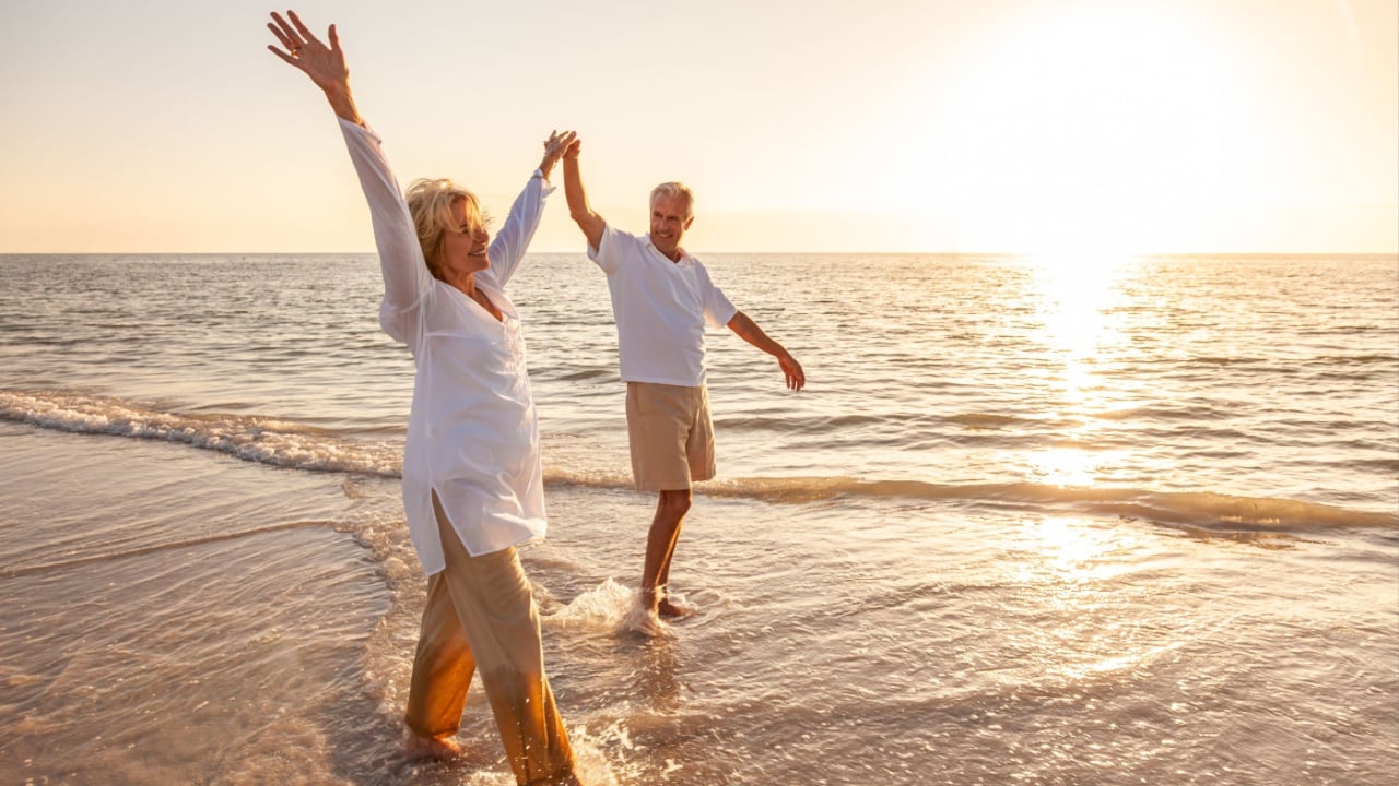 Happy senior man and woman old retired couple walking and holding hands arms raised on a beach at sunset