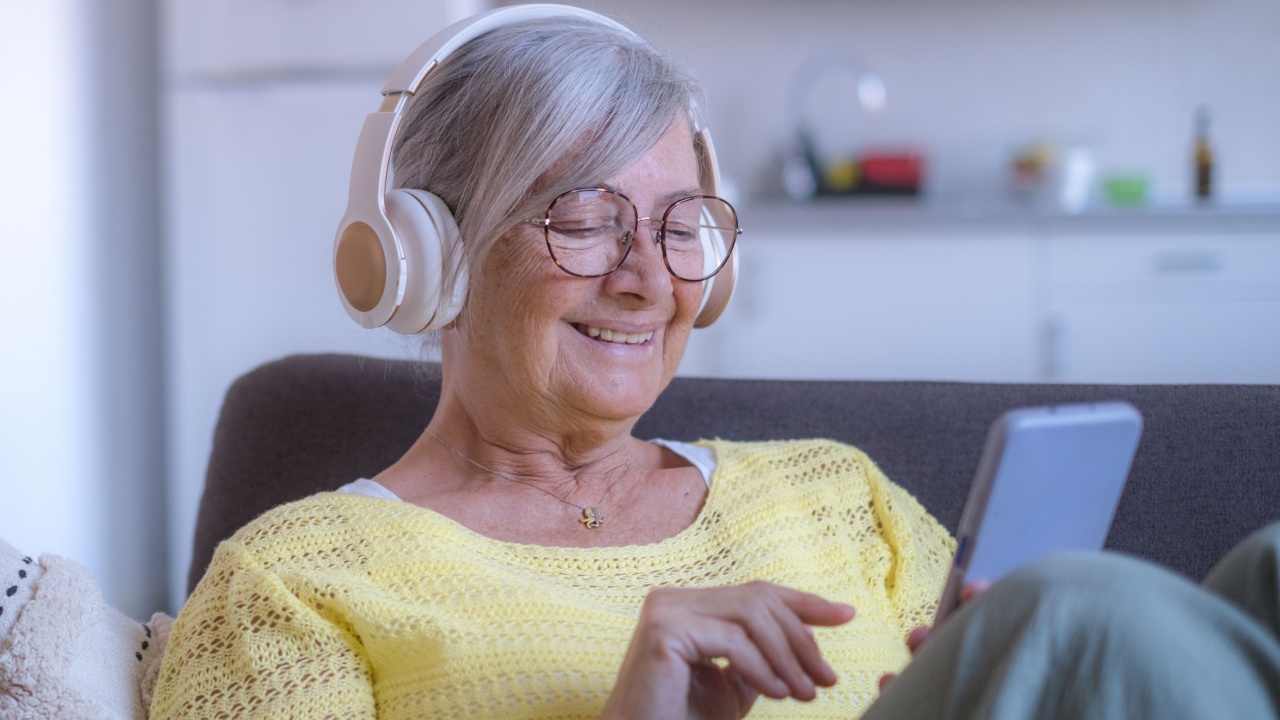 Happy senior retired woman sitting on sofa wearing headphones, using mobile phone technology, enjoying carefree moments communication chat video call with family or friends