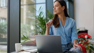 Young professional woman is focused on her laptop work at a bright modern office desk by the window