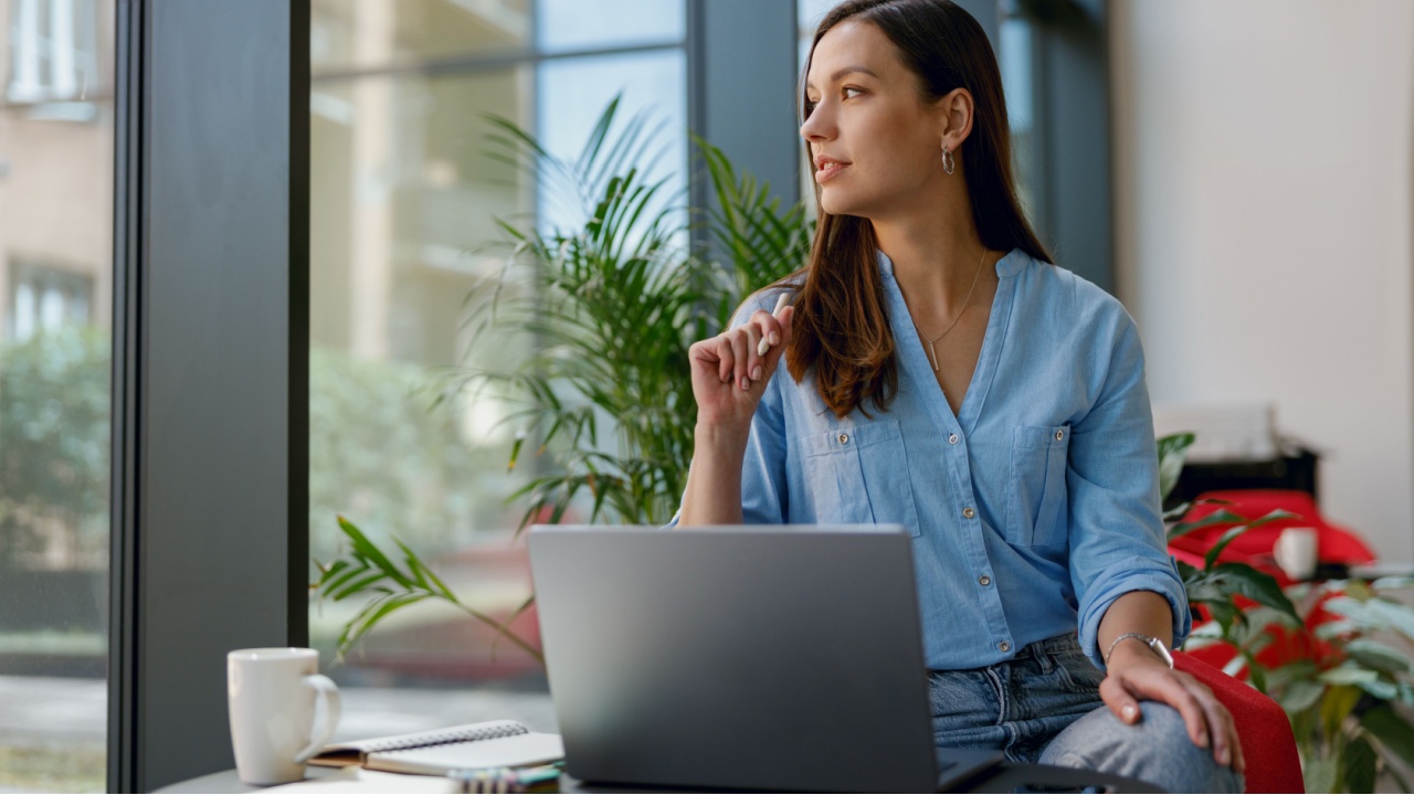Young professional woman is focused on her laptop work at a bright modern office desk by the window