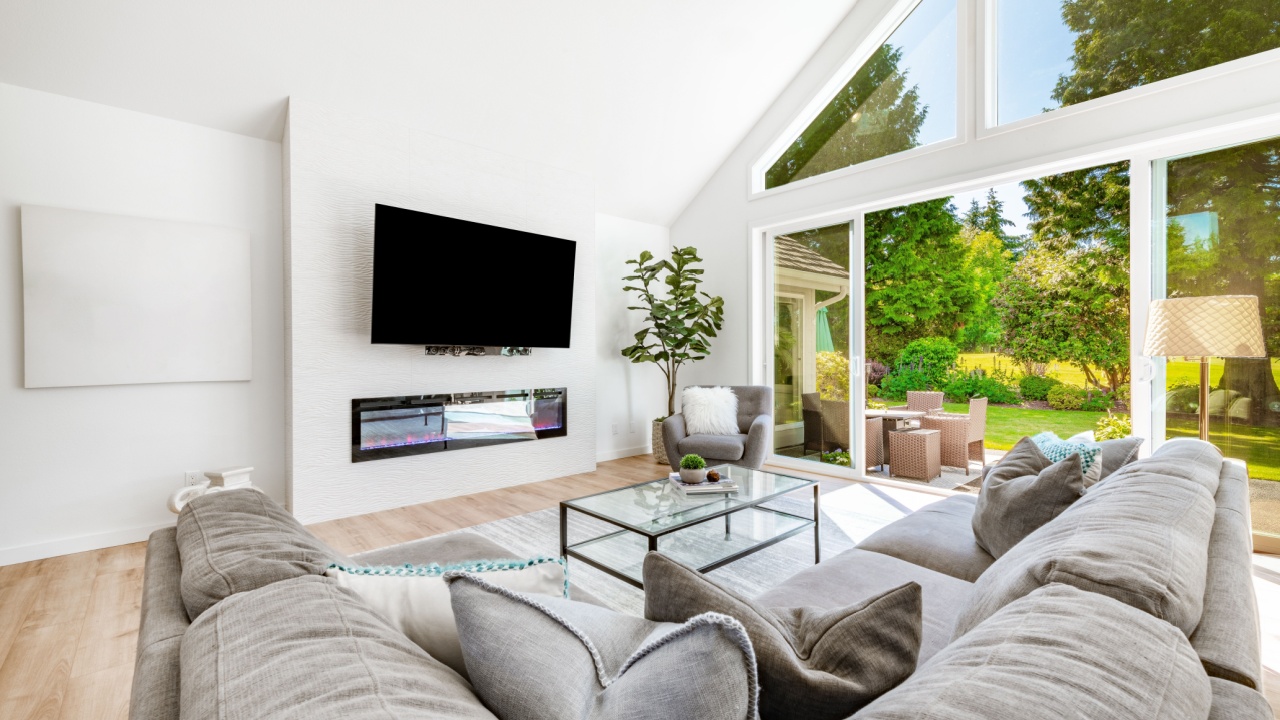 Large open concept living room with multiple expansive windows with view to garden landscaping comfortable furniture tv recliner electric fireplace hardwood floor white interior houseplant area rug