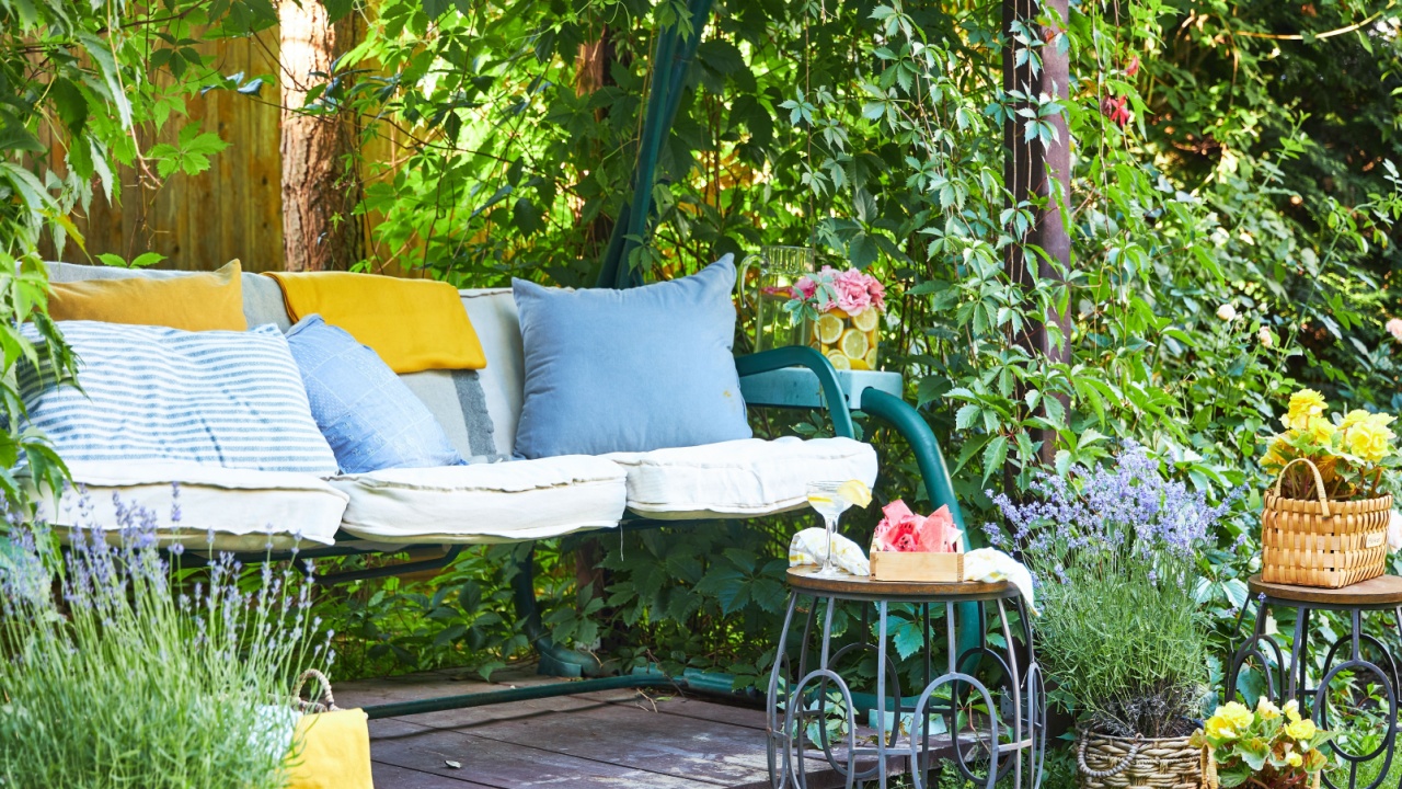 A cozy place to relax in the garden courtyard in a yellow and blue palette, garden swing curled with vines, with soft cushions, flowers in cachepots and watermelon slices on the table