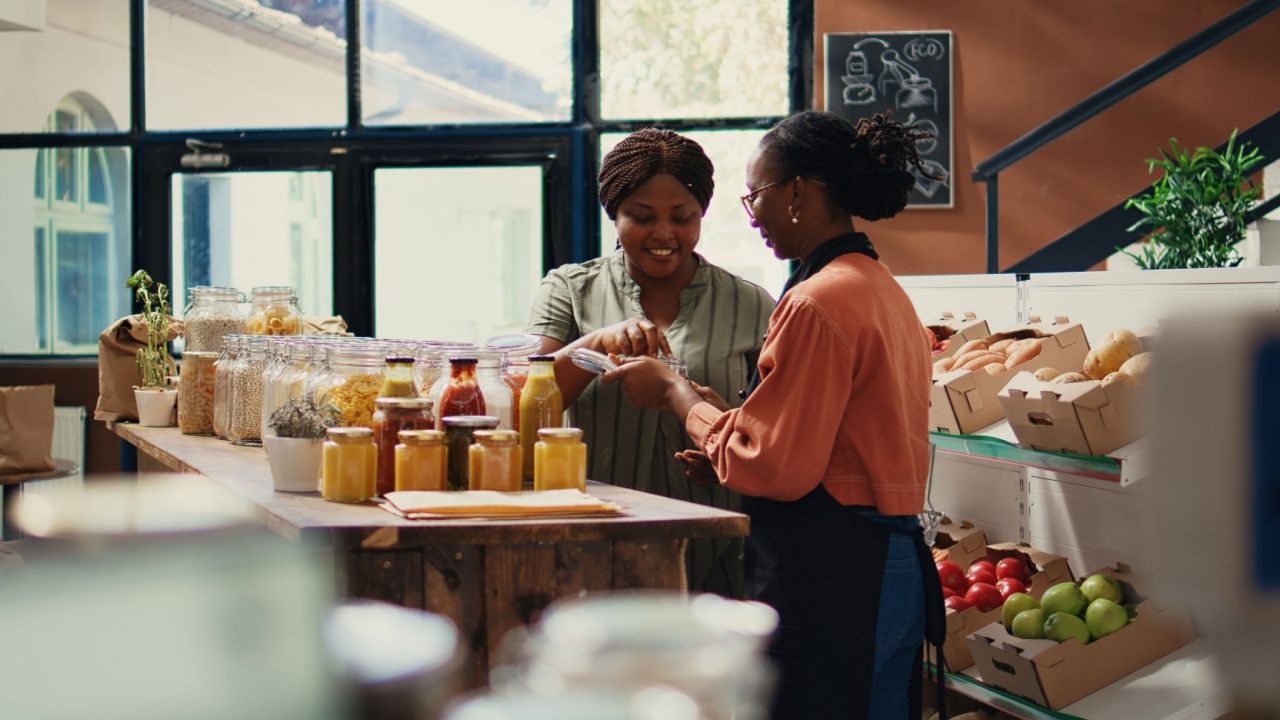 Retailer gives food samples to her customer in zero waste store, promoting new organic recipe for homemade snacks. Merchant recommending vegan alternatives for client in supermarket. Handheld shot.