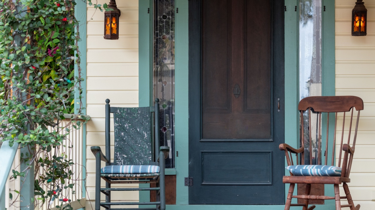 Two antique wooden rocking chairs on the front porch of a country style house. The door is wooden with green trim and the exterior wall is yellow. The patio furniture has cushions on the seats.