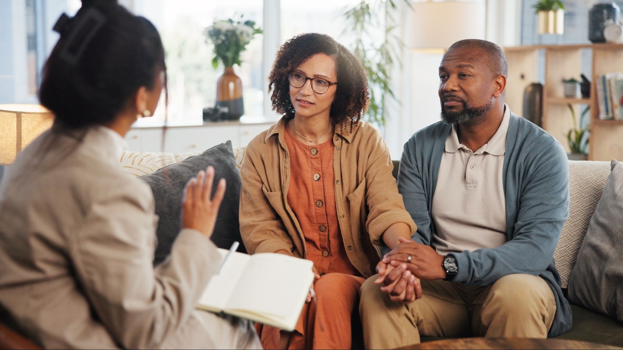 Couple, therapy and holding hands with discussion on sofa for guidance, communication and advice. Consultation, service and therapist with book in office for marriage counseling, support and progress