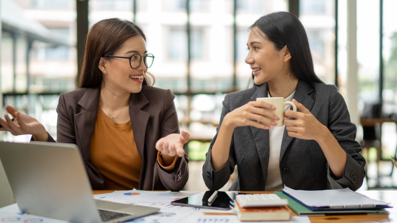 Two women are sitting at a desk, one of them is holding a cup of coffee. They are smiling and talking to each other. The scene suggests a friendly and relaxed atmosphere