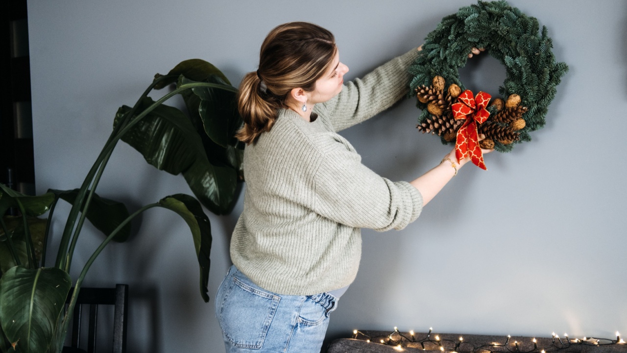 Woman hanging handmade Christmas wreath with pinecones and red ribbon. DIY holiday decor adds a personal touch to festive home decorating.