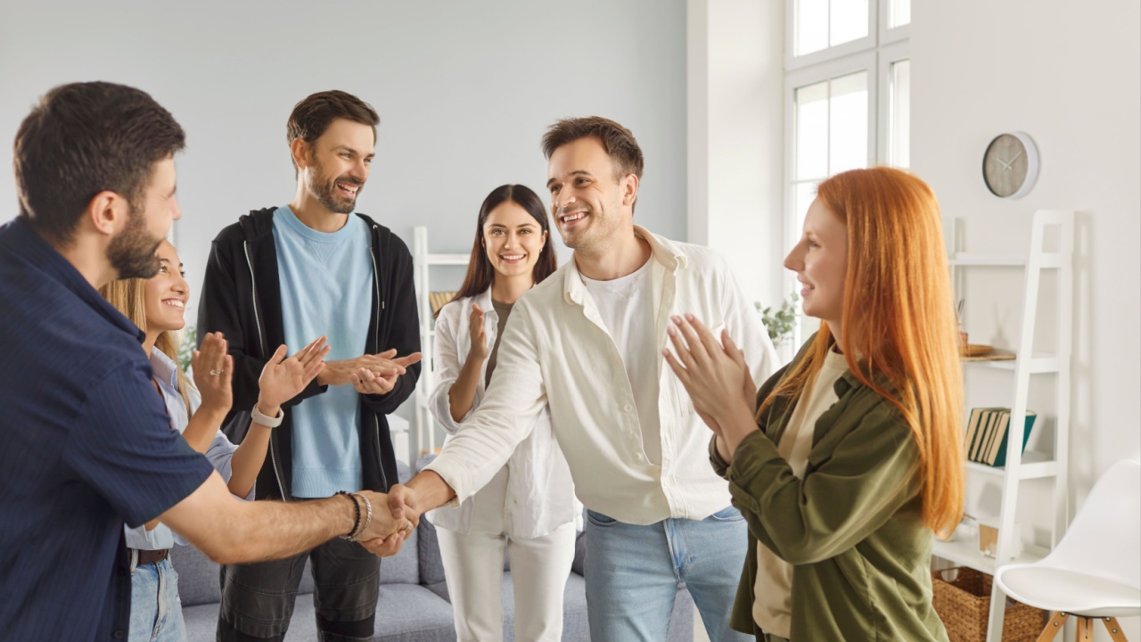 Group of a happy young friends men and women meeting at home standing in circle and shaking hands indoors greeting each other gathering indoors. Friendship, party, people and home leisure concept.