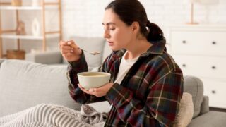 Sick young woman with plaid eating soup on sofa at home