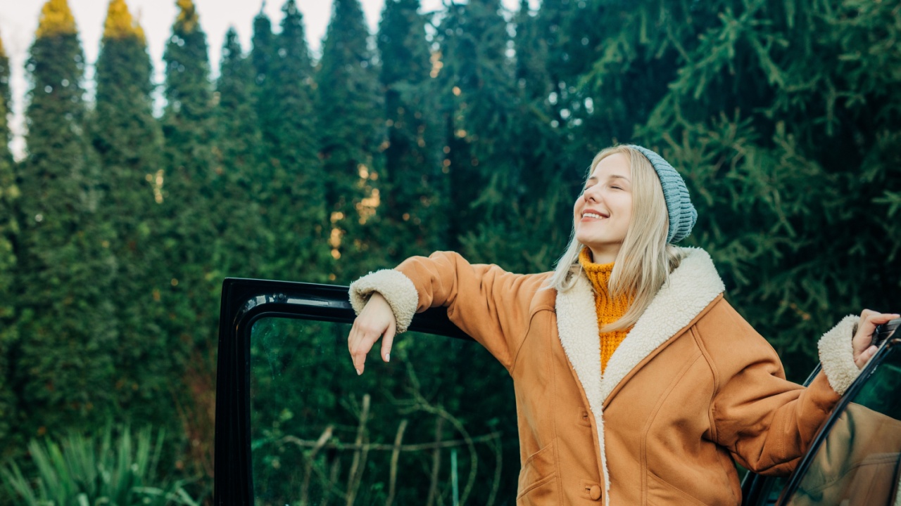 A blonde girl in a brown shearling jacket and a blue hat standing next to the door of a car with pine trees in the background