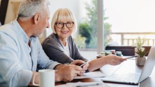 Smiling elderly husband and wife sit at table in living room manage household document pay bills on laptop together, mature 60s couple spouses read bank contract use online system on computer