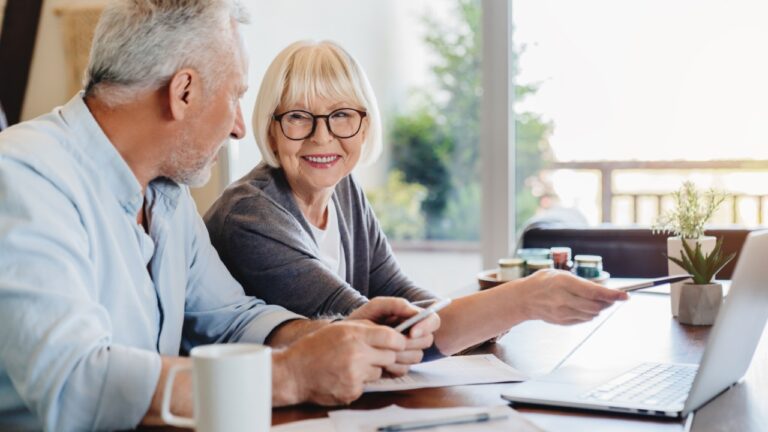 Smiling elderly husband and wife sit at table in living room manage household document pay bills on laptop together, mature 60s couple spouses read bank contract use online system on computer