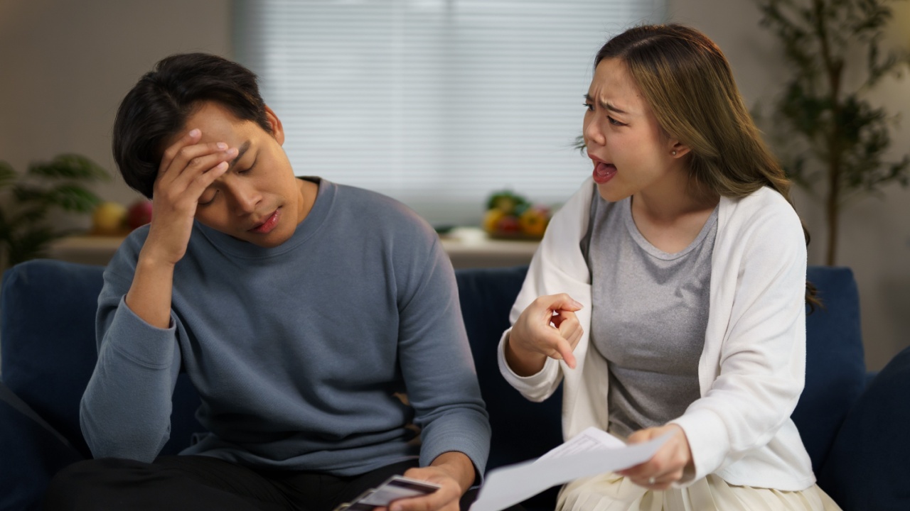 Young woman arguing at her husband about excessive credit card expenses, showing him bills and blaming him for overspending, while he is touching his head, stressed and overwhelmed by the situation
