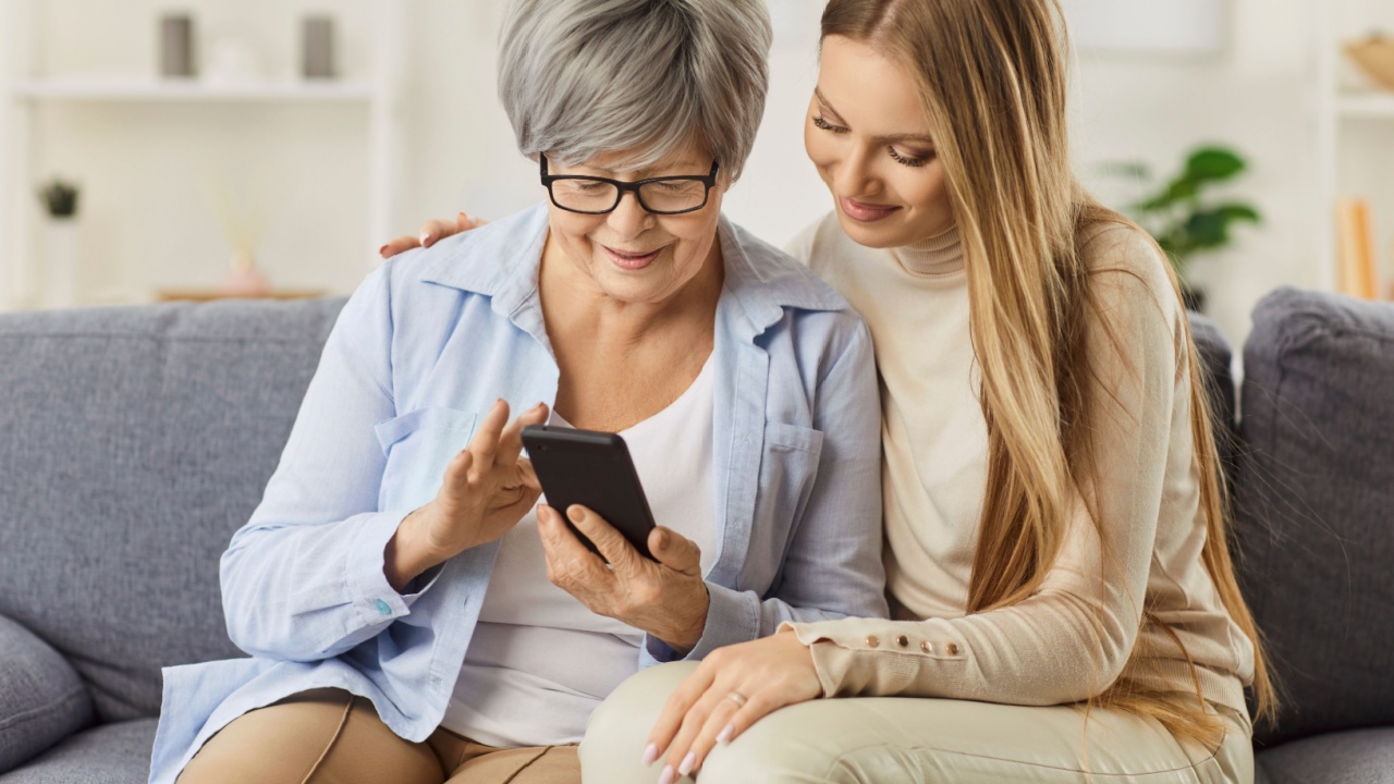 Helping senior mother with phone spp, adult daughter, affectionate young granddaughter and elderly grandma sitting close at home, caring happy family enjoying tender moments showing love and care