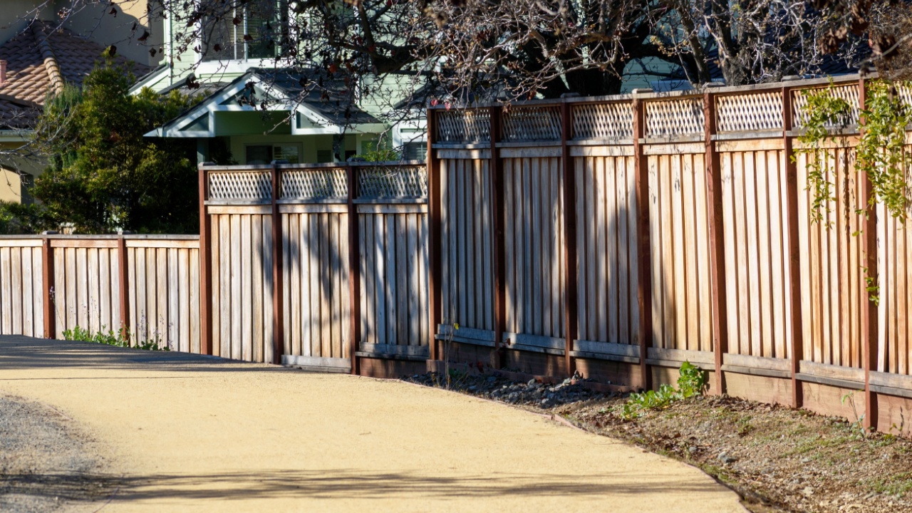 A wooden fence with lattice tops lines a curved dirt pathway, bordered by sparse vegetation, in a suburban neighborhood with visible house roofs and windows