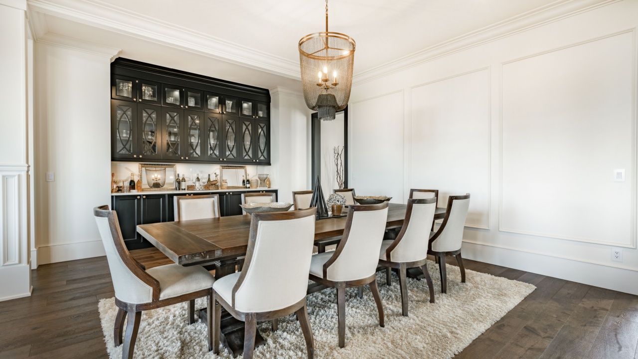 large luxury dining room home interior table seating ten chairs hardwood flooring area rug bar with bottles of alcohol liquor chandelier wainscotting panelled white walls columns beams elegant