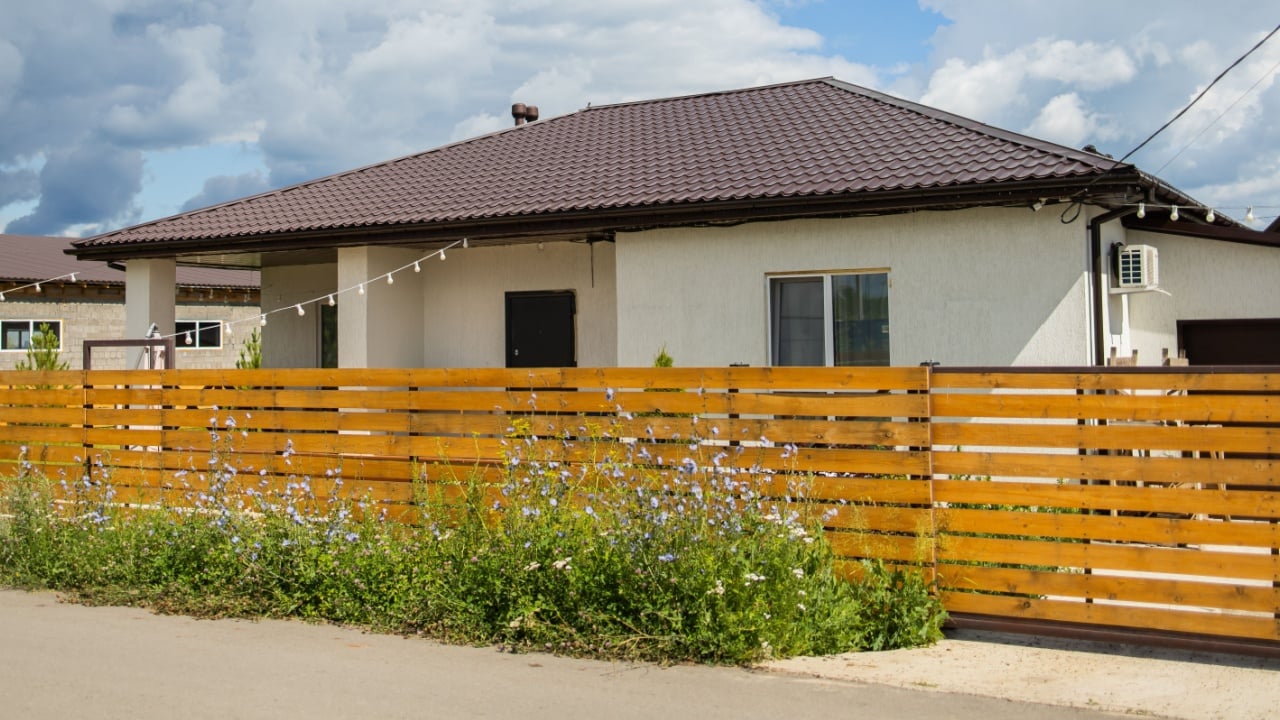 One-story house behind wooden fence and blooming wildflowers. Suburban privacy and residential space integration with natural elements.
