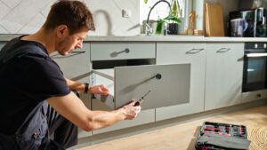 Man assembling kitchen drawer in modern cabinet using screwdriver. Handyman installing and adjusting drawer slide mechanism in contemporary kitchen. Furniture installation