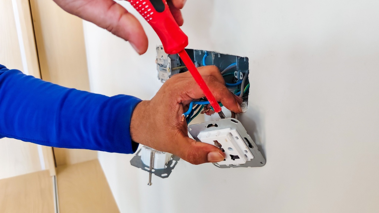 An electrician using a screwdriver to install or repair a wall-mounted electrical outlet, with wires visible inside the wall box.