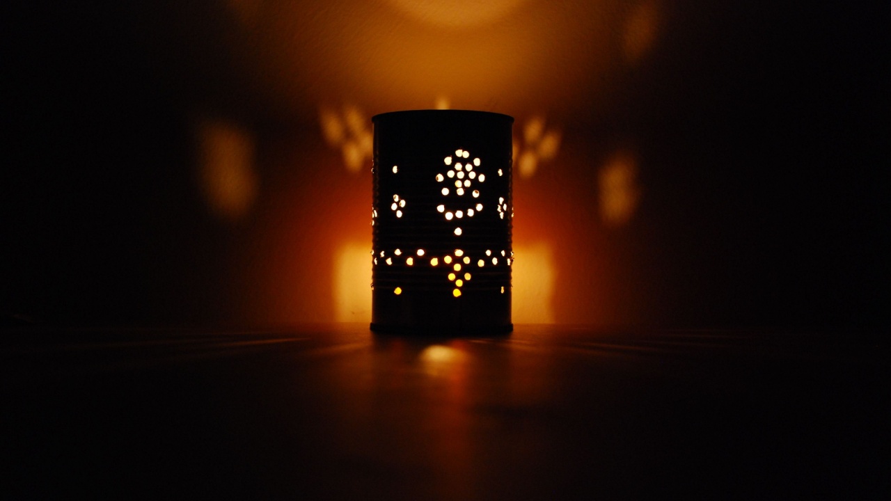 A handmade lantern created from a recycled tin can, lit by a candle inside, casting artistic shadows on the wall and table in a dimly lit room. The warm glow creates a cozy and intimate atmosphere
