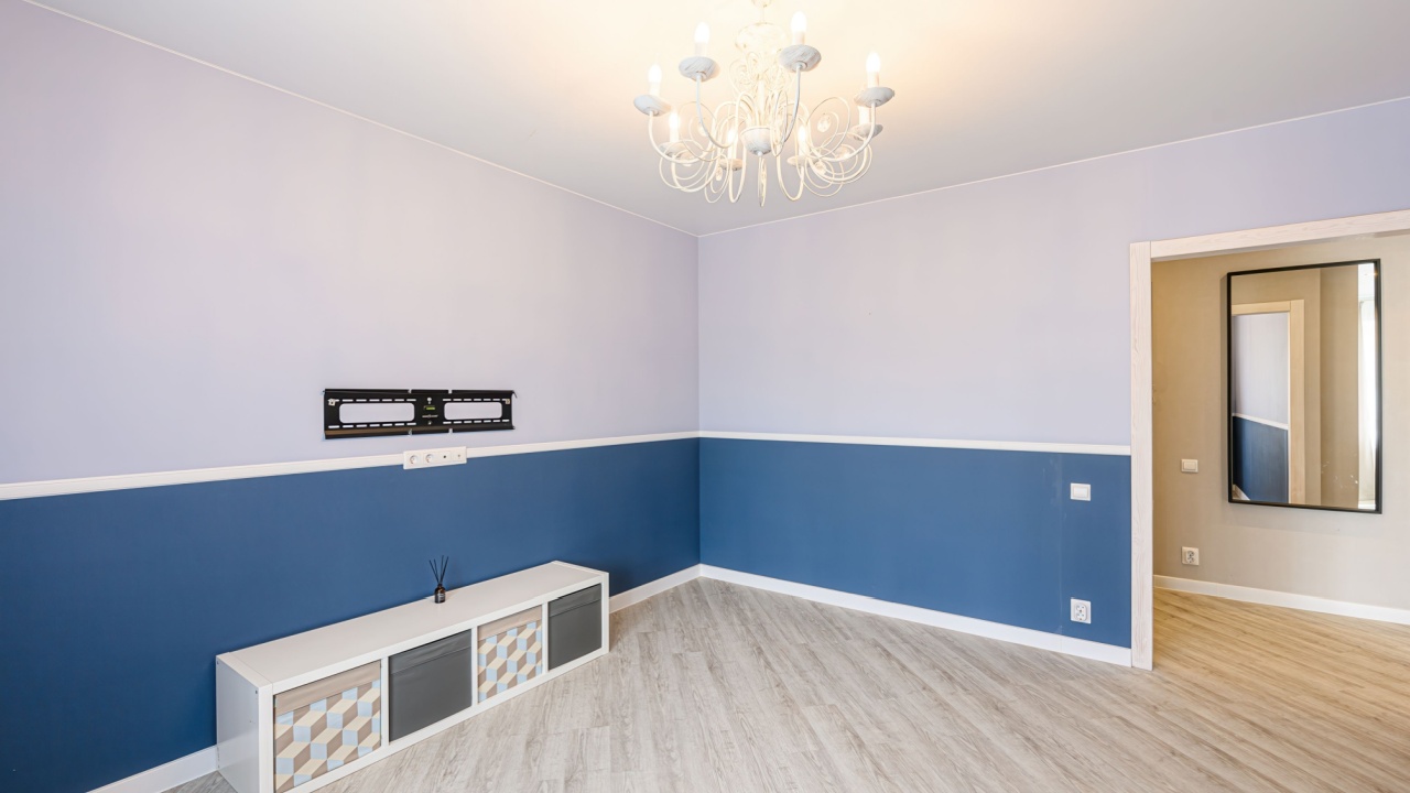 bright, empty room with two-tone walls (blue and lavender), light wood floors, a white shelf unit, and a white chandelier. A doorway leads to another room with a mirror